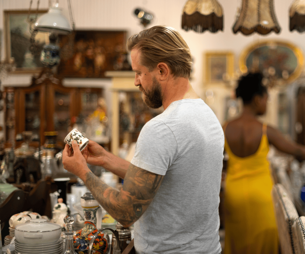 Man examining an antique porcelain cup in a vintage store filled with collectibles, decorative ceramics, and ornate furniture.