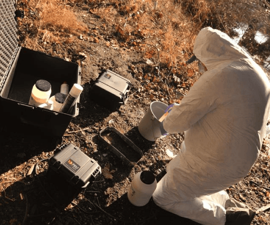 Scientist in protective suit collecting soil or water samples during environmental field testing, surrounded by sample containers and hard cases. Why this alt text works: Describes the action and setting clearly: Highlights “scientist,” “protective suit,” “soil or water samples,” and “environmental field testing”, key for accessibility and SEO relevance. SEO-friendly keywords: Includes terms like “environmental sampling,” “field testing,” “protective suit,” and “sample containers”, targeting environmental science, contamination studies, and field research audiences. Concise and accessible: 118 characters, within the 125-character limit recommended for screen readers. If you want to highlight specific fields (e.g., hazardous material testing, soil contamination, water quality monitoring), I can tailor the alt text even more!